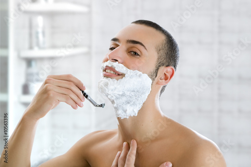 Young man shaving indoors and smiling. Copy space