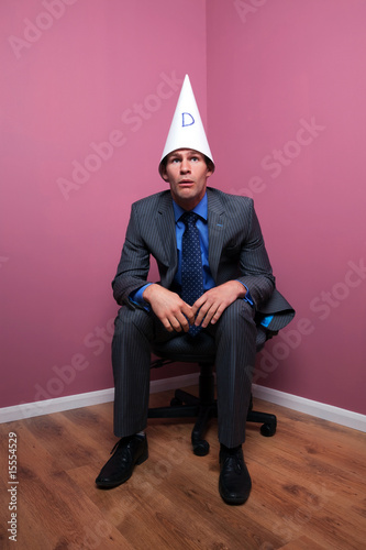 Businessman sat in corner wearing a dunce hat
