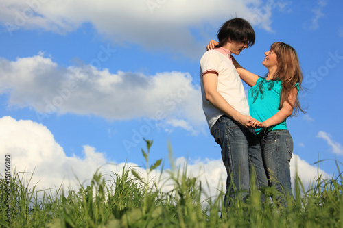 young pair holds each other in grass against sky