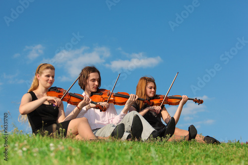 Three violinists sit and play on grass against sky