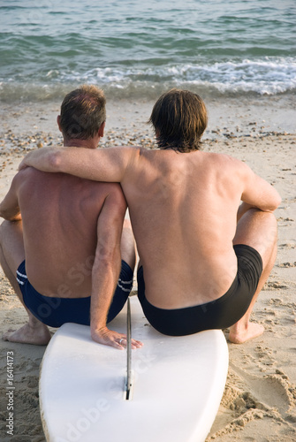 two gay men sitting on surfboard cuddling at the beach.