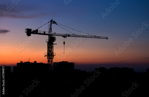 Tower crane silhouette at sunset over an urban construction site and city skyline. Industrial architecture, engineering, development and infrastructure project against orange dusk sky.