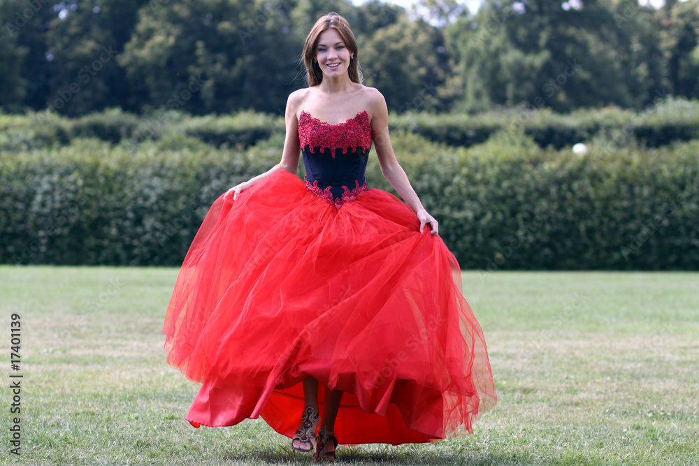 Fototapeta premium Smiling woman in a red dress walking on meadow in park