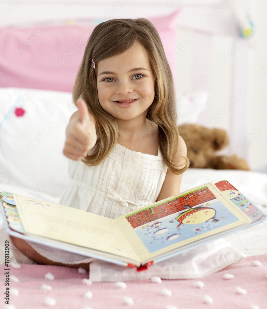 Happy little girl reading in bed with thumb up Happy little girl reading in bed with thumb up