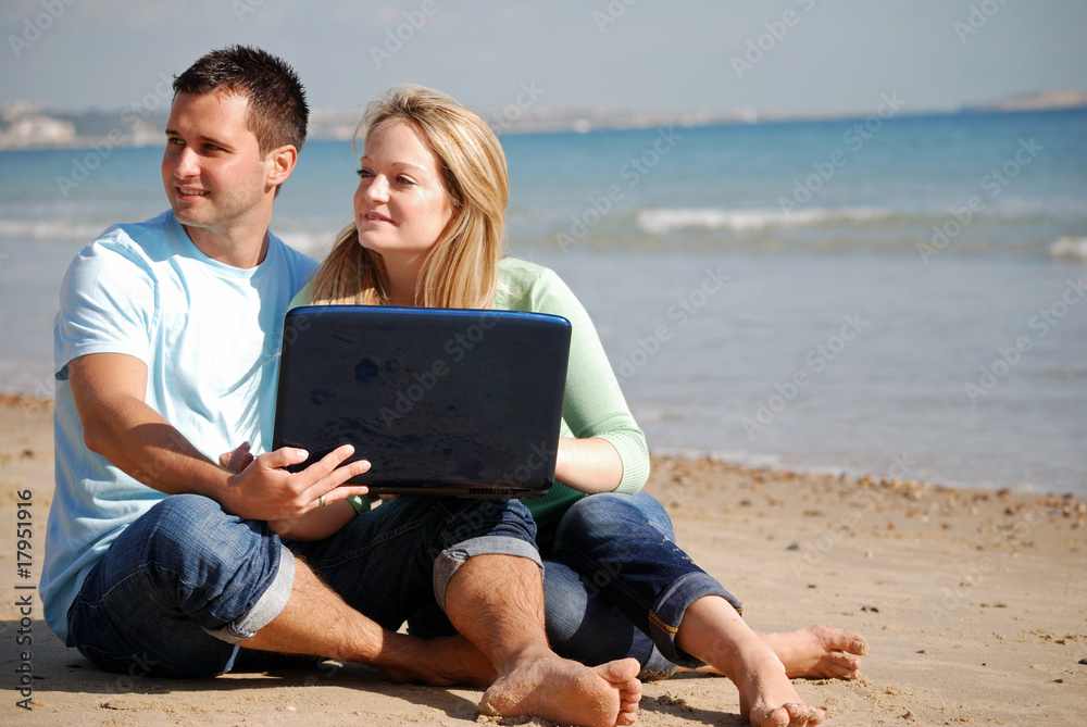 Couple sat on beach together with laptop