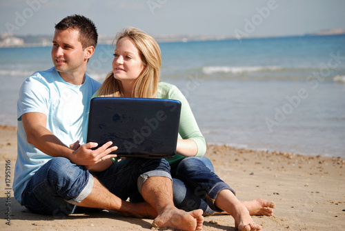 Couple sat on beach together with laptop