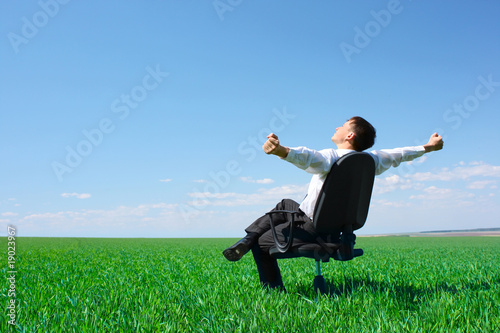 A young man with raised hands sitting in char on green meadow and looking to a sky with clouds