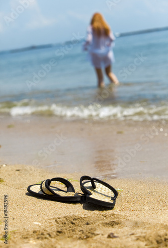 Beach slippers on sand and girl in ocean out of focus