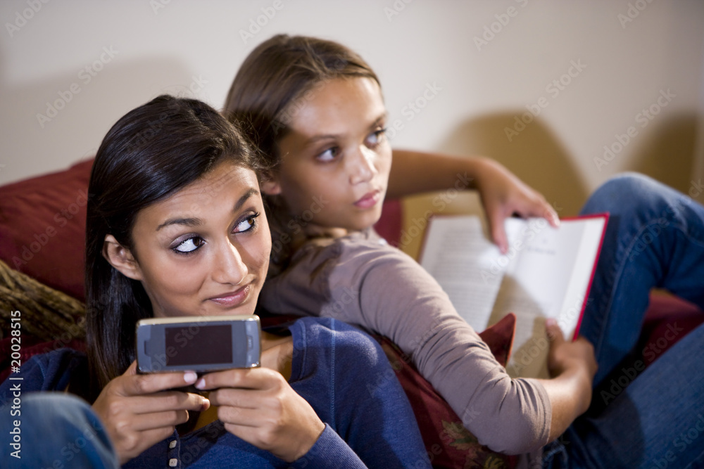 Girls reclining on sofa look up from texting and reading book Girls reclining on sofa look up from texting and reading book