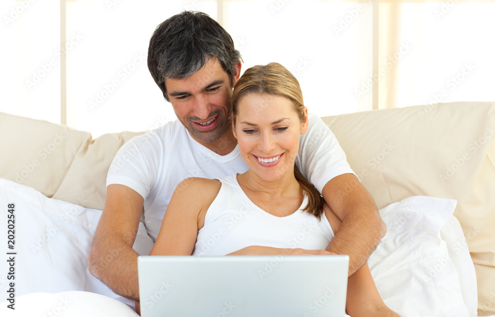 Fototapeta premium Enamoured couple using a laptop lying in the bed