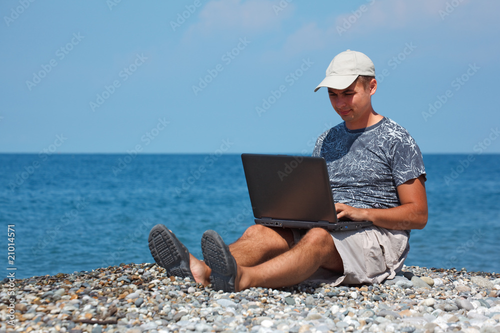 Man in cap sitting on beach with laptop on his knees