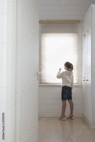 young girl peering through blinds standing at window full length