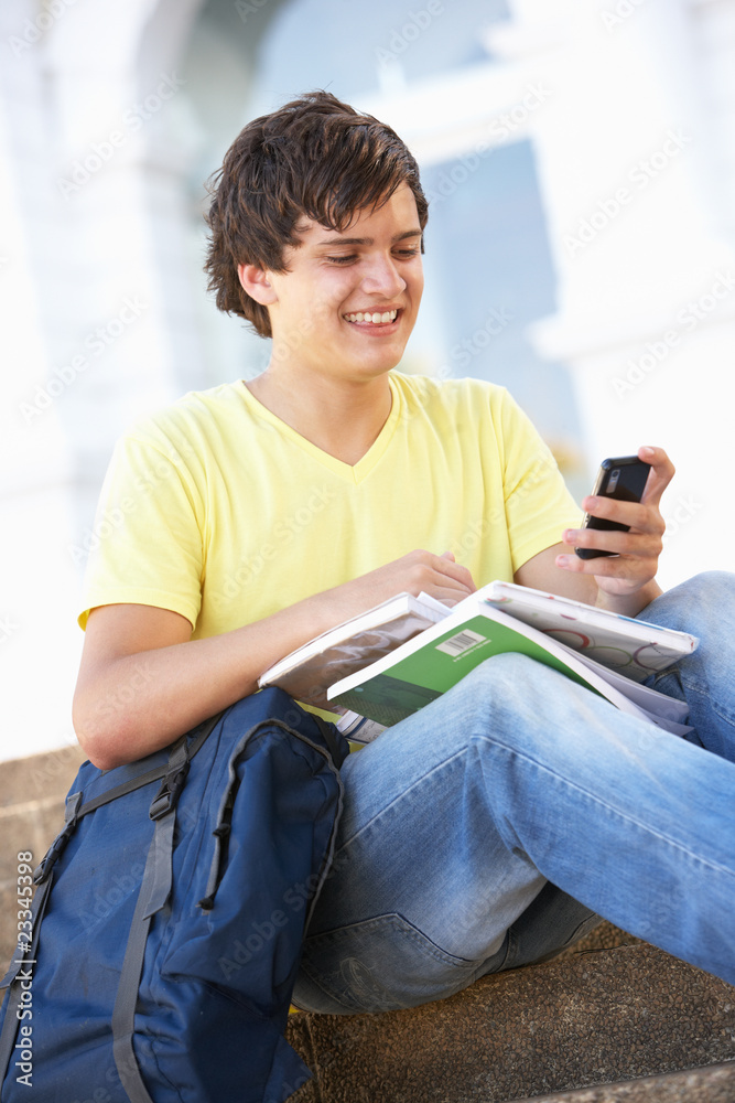 Male Teenage Student Sitting Outside On College Steps Using Mobi