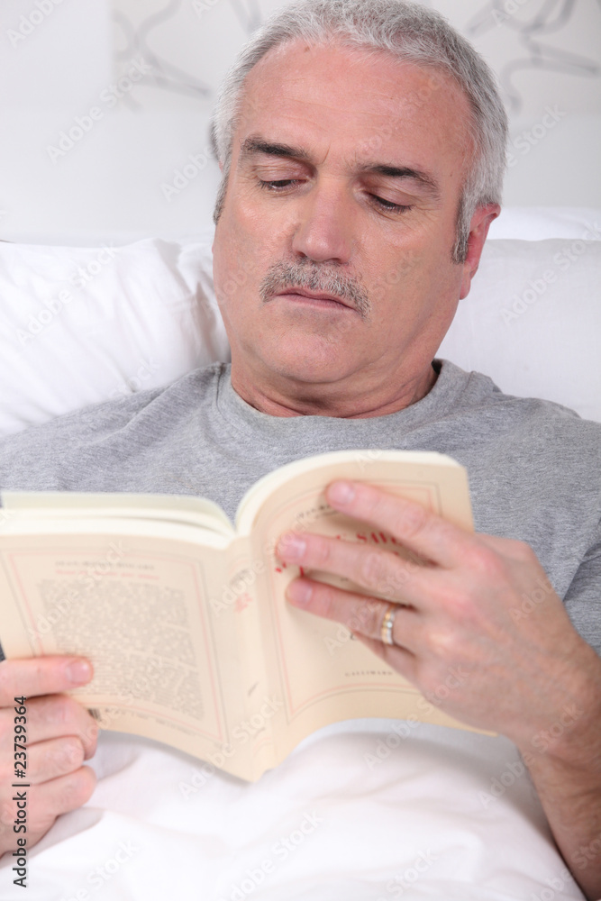 Homme senior lisant un livre allongé dans un lit Homme senior lisant un livre allongé dans un lit