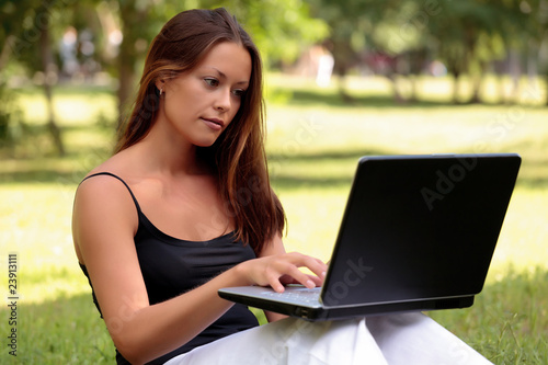 Beautiful young woman sits on a grass in a park with the laptop.