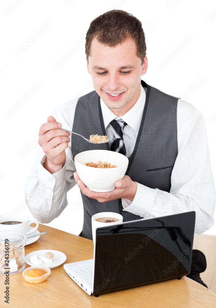 Happy businessman eating cereals looking at his laptop Happy businessman eating cereals looking at his laptop