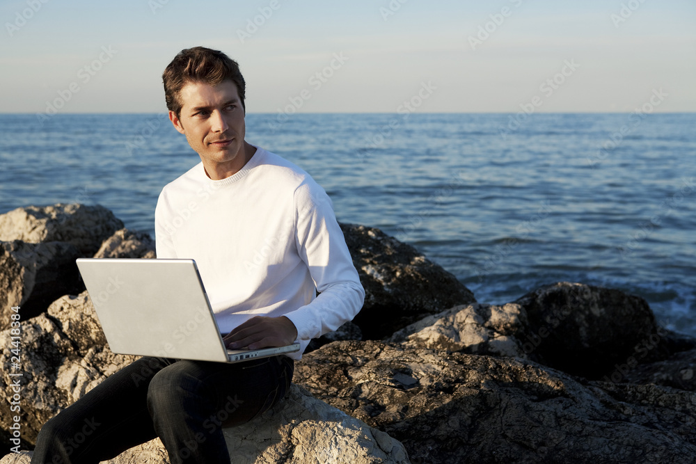 Young handsome man using laptop at beach, ocean in background