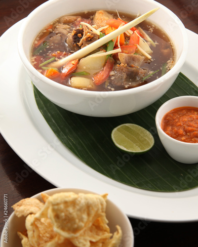 A close-up photograph of Indonesian Sop Buntut served in a bowl with a banana leaf used as an underliner, accompanied by a side of sambal. This dish showcases the hearty oxtail soup enriched with trad photo