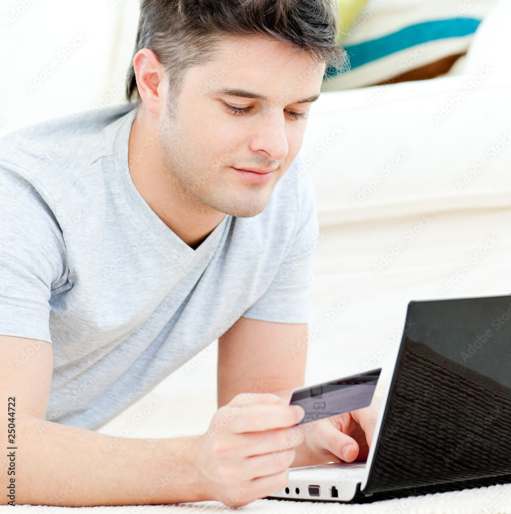 Positive young man holding a card using his laptop on the floor Positive young man holding a card using his laptop on the floor