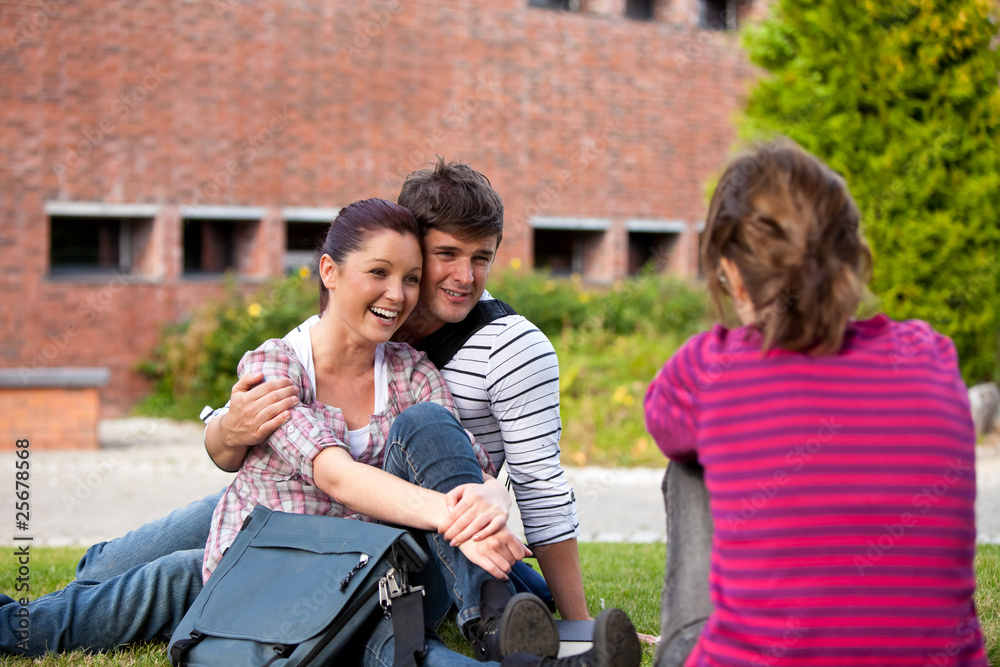 Fototapeta premium Happy couple of students sitting on grass talking with a female