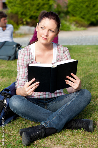 Bright female student reading a book sitting on the grass