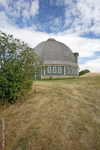A nice round barn sits partly behind a green bush.