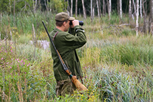 Hunter with rifle and binoculars in a forest`s swamp.