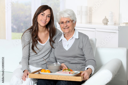 Elderly woman and home carer sitting in sofa with lunch tray