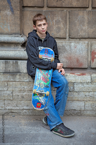 The teenager costs at a wall with a skateboard and has a rest.