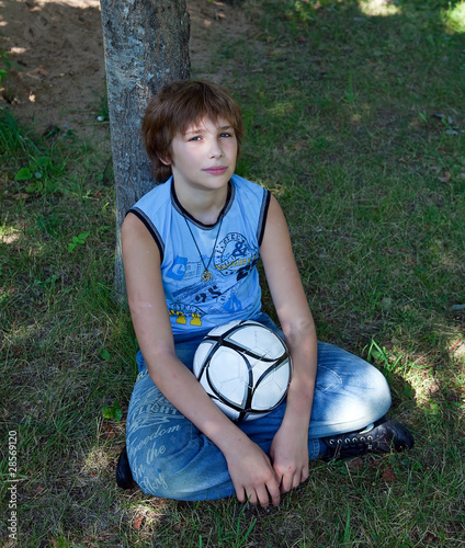 The teenager sits under a tree with a football.