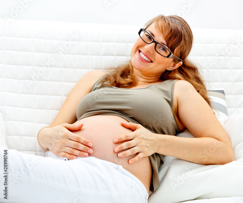 Smiling pregnant woman sitting on sofa and holding belly.