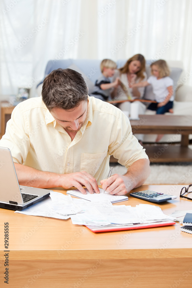 Man calculating his bills while his family are on the sofa Man calculating his bills while his family are on the sofa