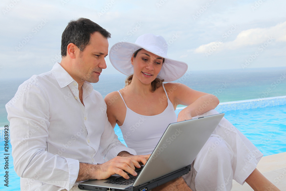 Couple using laptop computer by swimming-pool Couple using laptop computer by swimming-pool