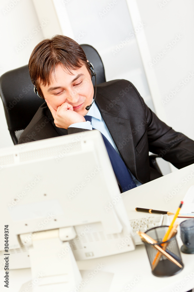 Smiling businessman sitting at desk and talking on headset . Smiling businessman sitting at desk and talking on headset .