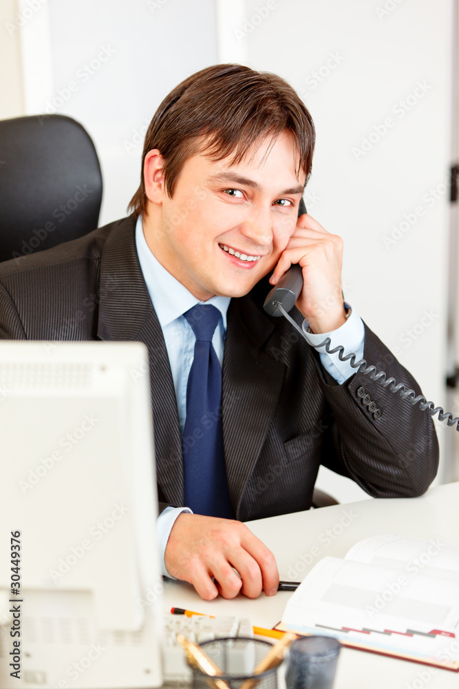 Friendly businessman sitting at desk and talking on phone. Friendly businessman sitting at desk and talking on phone.