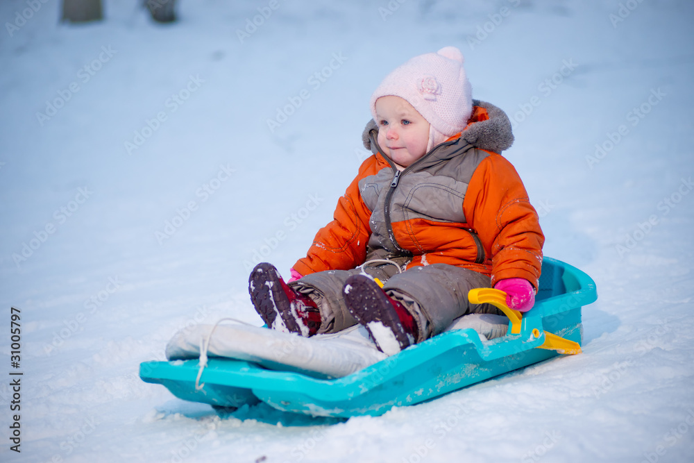 Adorable baby sliding on sleigh from hill in park and smile
