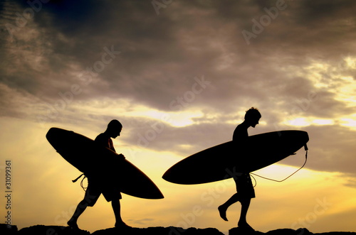 Fototapeta Naklejka Na Ścianę i Meble -  Two Surfers Carrying Their Boards Home At Sunset