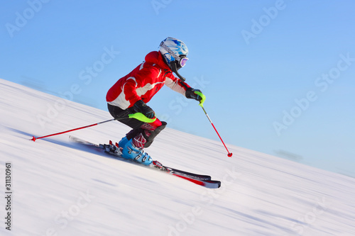 Skier in red jacket riding on hill. Motion blurred snow and clear blue sky