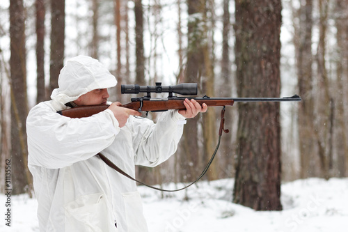 Sniper in white camouflage aiming with rifle at winter forest.