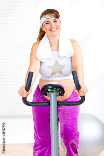 Smiling pregnant woman working out on static bike at home.