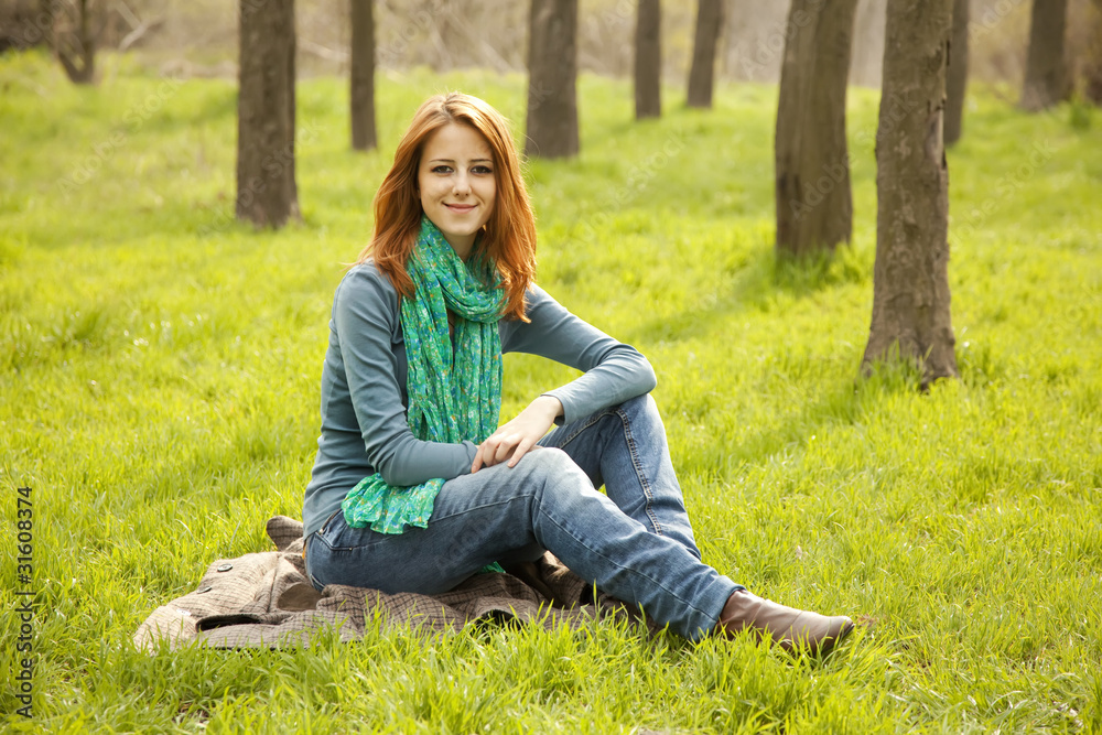 Obraz premium Beautiful red-haired girl sitting at green grass at park.