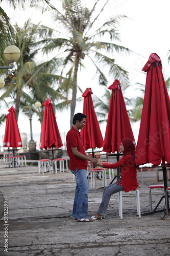 A cheerful Asian Muslim couple spending time together outdoors, dressed in matching red outfits. The man and woman are smiling and showing affection in a peaceful natural setting. This photo captures 