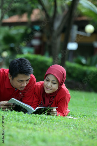 A cheerful Asian Muslim couple spending time together outdoors, dressed in matching red outfits. The man and woman are smiling and showing affection in a peaceful natural setting. This photo captures 