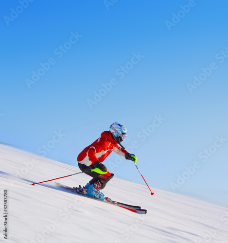 Skier in red jacket riding on hill. Motion blurred snow and clear blue sky