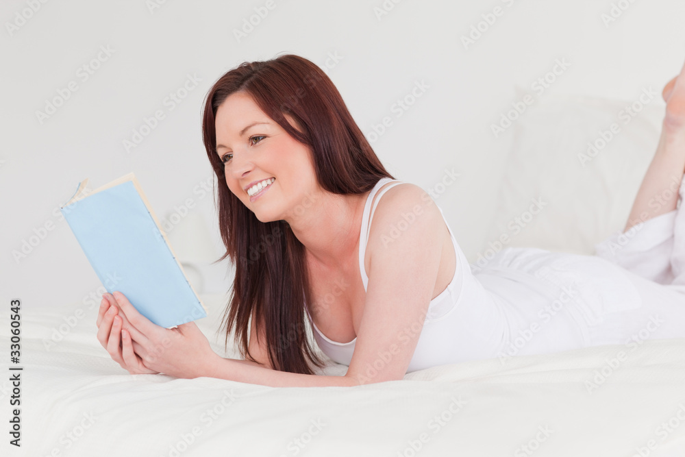 Gorgeous red-haired woman reading a book while lying on her bed