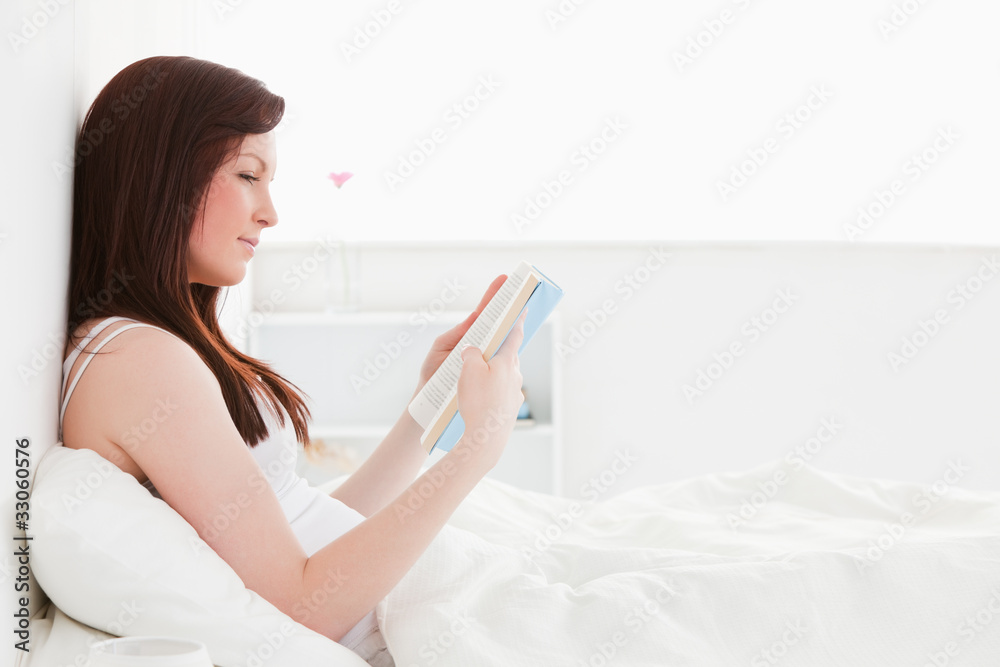 Pretty red-haired female reading a book while sitting on her bed Pretty red-haired female reading a book while sitting on her bed