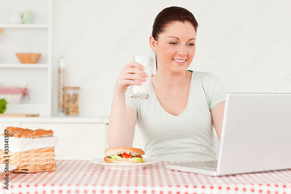 Smiling woman relaxing on her laptop while drinking a glass of m Smiling woman relaxing on her laptop while drinking a glass of m
