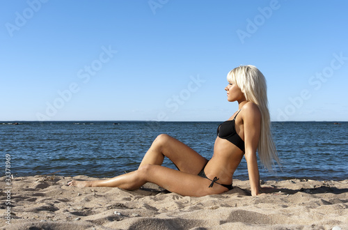 blonde girl in a black bathing suit sitting on shore blue sea