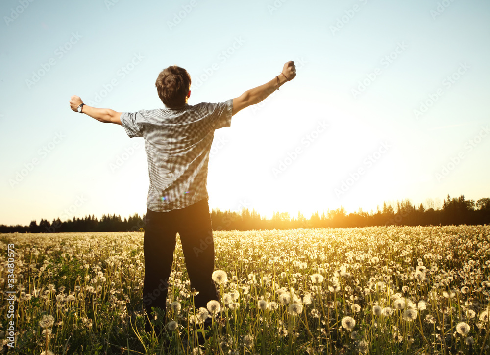 Young smiling man standing on a meadow with dandelions on a blue sky background