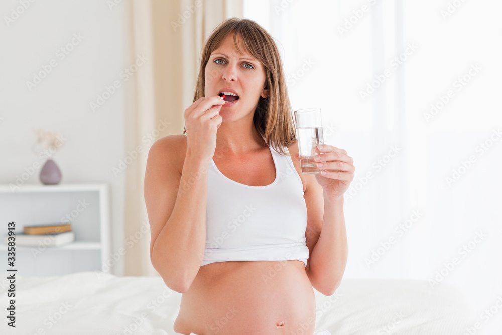 Attractive pregnant female taking a pill while sitting on a bed Attractive pregnant female taking a pill while sitting on a bed